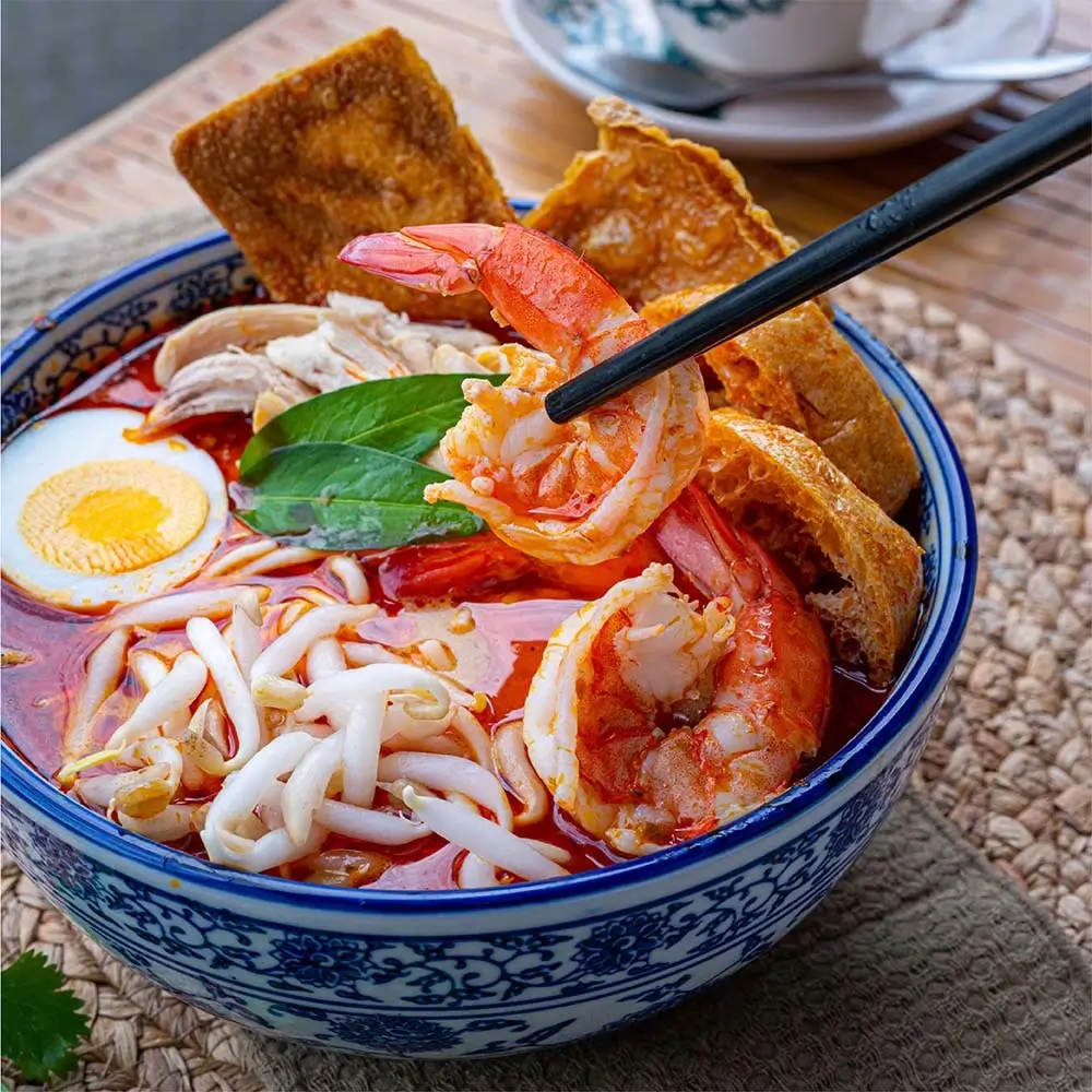 Malaysian prawn noodle soup with fresh shrimp, boiled egg, tofu puffs and bean sprouts in a blue ceramic bowl